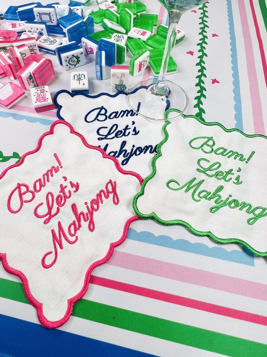 A trio of Mahjong cocktail napkins in bright pink, blue, and green, each with a matching embroidered border and text, laid out on a lively Mahjong-themed table setting.