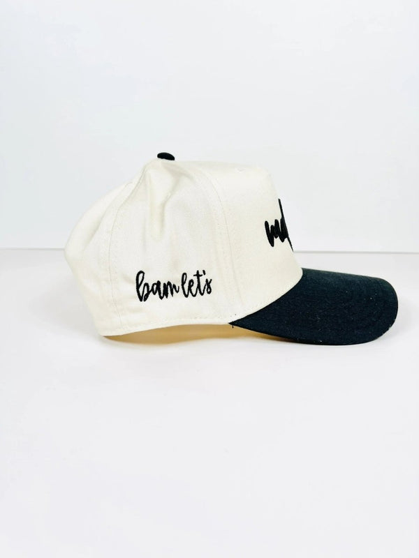 Side view of a cream and black Mahjong trucker hat with 'bam let's' embroidered in black cursive on the side. The cap has a structured front panel and a black brim for contrast.