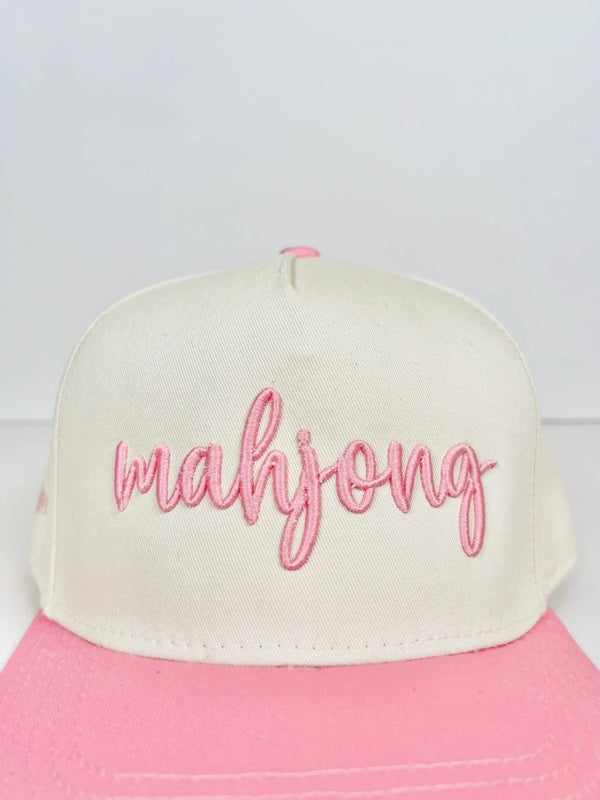Close-up of a cream and pink trucker hat with 'mahjong' embroidered in pink cursive on the front panel. The cap has a stylish, modern design with a curved brim.