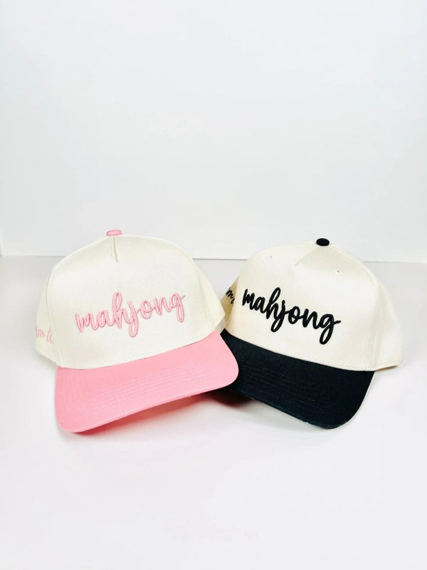 Two Mahjong trucker hats in cream with contrasting brims—one with a pink brim and pink embroidery, the other with a black brim and black embroidery. Both feature 'mahjong' in cursive on the front.